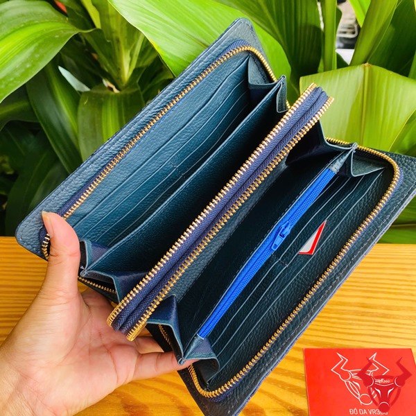 Sang Trọng Thời Trang: Ví Nam Nữ BIA4 Xanh Navy Đẳng Cấp Cao Cấp Tiện Dụng Hình ảnh ví da cá sấu xanh navy cầm tay, nền lá xanh, thẻ thương hiệu đỏ bên cạnh.