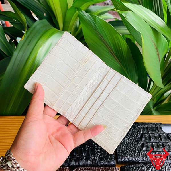 Da Bụng Cá Sấu VSAA8 Trắng Mộc – Ví Thời Trang Cao Cấp Đẳng Cấp Sang Trọng Hình ảnh cầm ví da bụng cá sấu trắng mộc VSAA8, nền gỗ với cây xanh và logo bò.