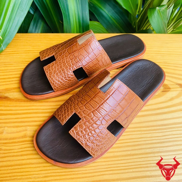 Thời trang dép cao cấp size 41, bền đẹp êm chân tối ưu. Đôi dép da cá sấu chữ H DJA8 màu nâu nhạt size 41 trên bàn gỗ với nền xanh.