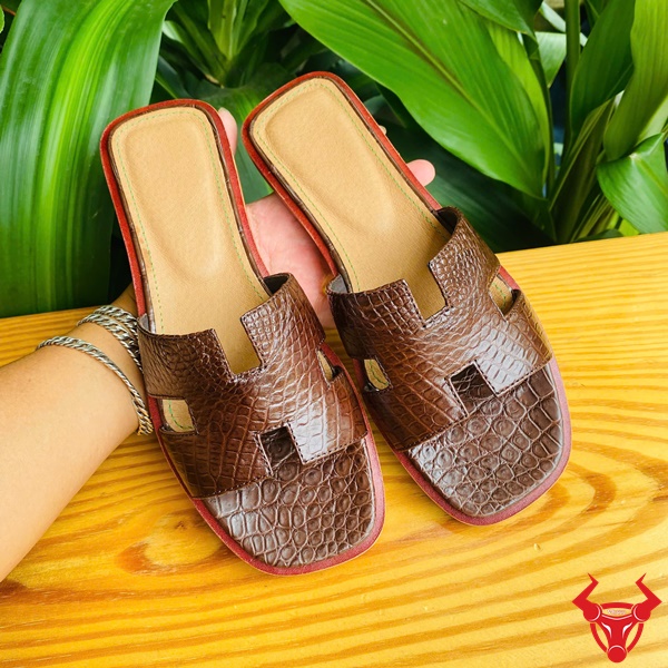 Cao cấp thời trang dép nữ, bền đẹp và êm chân vượt trội. Hình ảnh dép nữ DJBA9 da cá sấu chữ H màu nâu đất với nền lá xanh tươi.