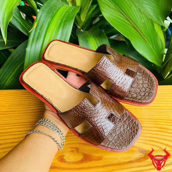 Thời trang dép nữ cao cấp, bền đẹp và êm chân tối ưu. Sản phẩm dép nữ DJBA9 da cá sấu chữ H màu nâu đất trong không gian tự nhiên.