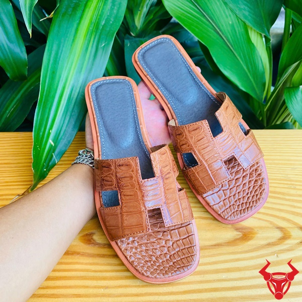 Dép da cá sấu cao cấp thời trang, bền đẹp êm chân vượt trội tự nhiên. Sản phẩm dép nữ DJBA9 da cá sấu chữ H màu nâu nhạt trong không gian tự nhiên xanh.