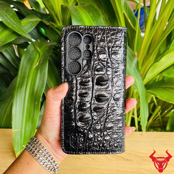 Đẳng Cấp Thời Trang Với Bao Da Cá Sấu Đen Samsung S25 Hình bao da Samsung S25 da cá sấu đen, cầm tay giữa nền lá xanh, đại diện cho sự sang trọng cao cấp.