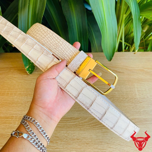 Đẳng Cấp Lịch Lãm Với Thắt Lưng Cá Sấu Bạch Tạng Thời Trang Hình thắt lưng da cá sấu bạch tạng với khóa vàng kim loại, cầm trên tay giữa nền thực vật xanh, tượng trưng cho sự sang trọng.