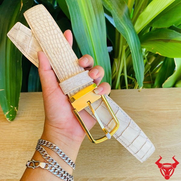 Thắt Lưng Cá Sấu Bạch Tạng - Biểu Tượng Cao Cấp Cho Quý Ông Ảnh thắt lưng da bụng cá sấu bạch tạng với khóa kim lấp lánh, cầm tay trước nền lá cây, biểu tượng phong cách cao cấp.