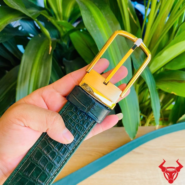 Phong Cách Nam Tính Từ Thắt Lưng Cá Sấu Xanh Lá Lịch Lãm Hình thắt lưng da cá sấu xanh lá liền mạch với khóa vàng kim, được cầm tay trước nền lá xanh, thể hiện gu thời trang đẳng cấp.