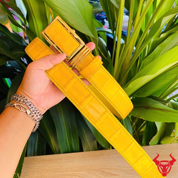 Đẳng Cấp Lịch Lãm Với Thắt Lưng Cá Sấu Vàng Thời Trang Hình thắt lưng da cá sấu vàng với khóa kim lấp lánh, cầm trên tay giữa nền thực vật xanh, tượng trưng sang trọng.