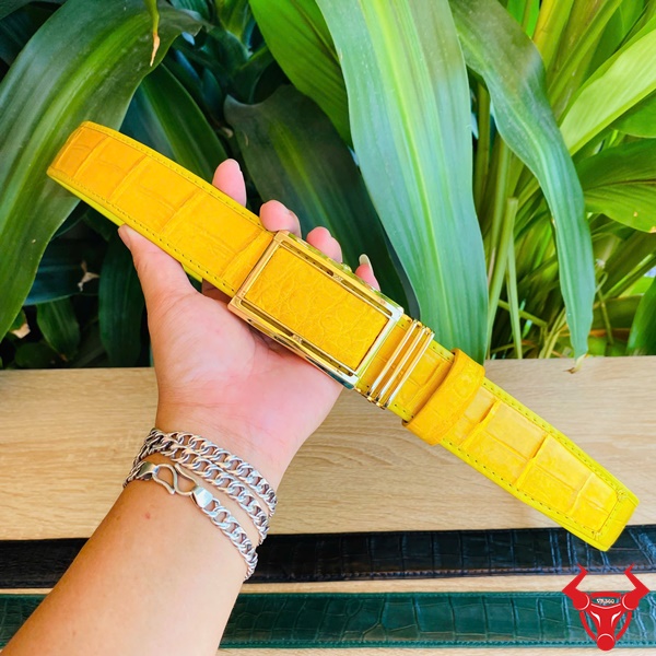 Phong Cách Nam Tính Từ Thắt Lưng Cá Sấu Vàng Lịch Lãm Thắt lưng da bụng cá sấu vàng với khóa vàng kim, cầm tay trước nền lá xanh, biểu tượng thời trang sang trọng.
